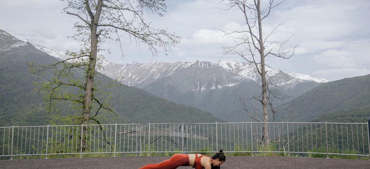 A woman doing a plank exercise on a mat outdoors with a scenic mountain view in the background.